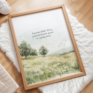 May include: A framed print with a watercolor landscape scene featuring trees, a field of wildflowers, and distant hills. The frame is made of light-colored wood. Text at the top reads: "Custom Bible Verse, your favourite quote or saying here."