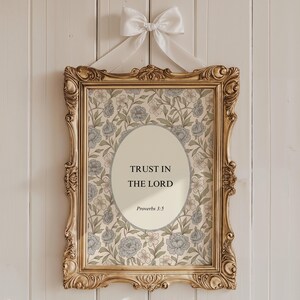 May include: A rectangular gold ornate frame containing a print with a floral pattern of blue flowers and green leaves. In the centre, an oval cream-coloured field displays the black text: TRUST IN THE LORD Proverbs 3:5. A white satin ribbon bow hangs above.