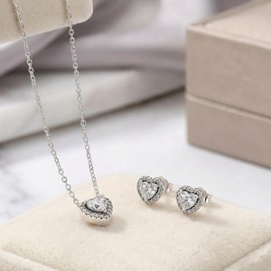 May include: A silver necklace featuring a heart-shaped pendant with a central stone surrounded by small clear crystals, accompanied by a matching pair of heart-shaped stud earrings, each with a central stone and a border of small clear crystals.