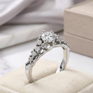 May include: A silver-coloured ring featuring a large, round, brilliant-cut centre stone held by a prong setting. The band is adorned with a series of smaller, round-cut stones that extend along the shoulders, creating a cluster effect on the metal band.