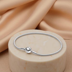 May include: A silver bracelet with a snake chain design. The bracelet has a round clasp with the letters "OP" and a small gemstone. The bracelet is displayed on a beige cylindrical stand against a neutral background.