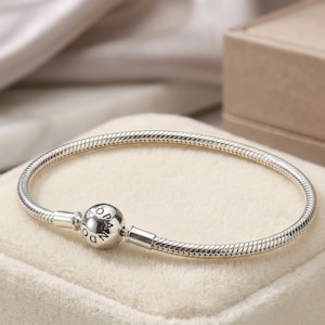 May include: A silver snake chain bracelet featuring a round barrel clasp. The clasp is engraved with the word PANDORA and a small crown symbol above the letter O. The bracelet has a smooth, flexible, and metallic finish with a polished surface.
