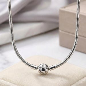 May include: A silver snake chain necklace featuring a spherical clasp. The round clasp is engraved with the text PANDORA and includes a small crown symbol above the letter O. The chain has a smooth, flexible, and segmented metallic texture.