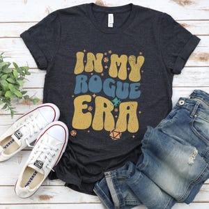 Könnte beinhalten: Ein dunkelgraues T-Shirt mit dem Text "In My Rogue Era" in einer Retro-Schriftart in verschiedenen Farben. Der Text ist von kleinen weißen Sternen und Würfeln umgeben.