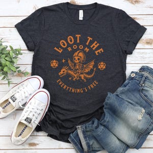 May include: Dark gray t-shirt with the text "LOOT THE ROOM EVERYTHING'S FREE" in orange. The graphic features a skeleton riding a dragon. White canvas sneakers and blue jeans are also in the image.