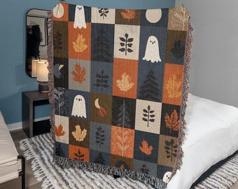 Puede incluir: Una manta tejida con un diseño de patchwork con fantasmas, hojas, árboles y lunas en tonos naranja, marrón, gris y crema. La manta tiene un borde con flecos y se exhibe en interiores.