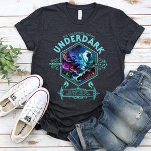 Może przedstawiać: Szary t-shirt z ciemnym melanżem z grafiką fantasy krajobrazu w niebiesko-zielonej sześciokątnej ramie. Tekst "The Shadowed Depths Underdark" znajduje się nad grafiką, a tekst "Forgotten Realms" po obu stronach grafiki. Tekst "Journey Below, Face The Unknown" znajduje się pod grafiką.
