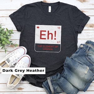May include: A dark grey heather t-shirt featuring a white square graphic resembling a periodic table element. The graphic displays the red text Eh!, a small red maple leaf, the numbers 18 and 67, and the text THE ELEMENT OF CANADA.