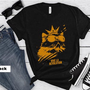 May include: A black short-sleeved t-shirt featuring a graphic print of a cat wearing a crown and sunglasses. The cat is rendered in orange, and the text "VIVE LA REVOLUTION" is printed in orange below the cat. The shirt has a crew neckline.