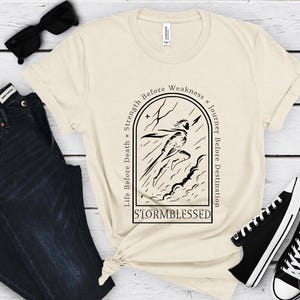 Puede incluir: Camiseta color crema con un gr&aacute;fico negro de una figura corriendo a trav&eacute;s de una tormenta, con la palabra "STORMBLESSED" debajo. La camiseta tiene las frases "Life Before Death, Strength Before Weakness, Journey Before Destination" alrededor del gr&aacute;fico. Gafas de sol negras, vaqueros azul oscuro y zapatillas negras tambi&eacute;n en la imagen.