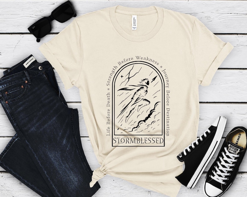 Puede incluir: Camiseta color crema con un gr&aacute;fico negro de una figura corriendo a trav&eacute;s de una tormenta, con la palabra "STORMBLESSED" debajo. La camiseta tiene las frases "Life Before Death, Strength Before Weakness, Journey Before Destination" alrededor del gr&aacute;fico. Gafas de sol negras, vaqueros azul oscuro y zapatillas negras tambi&eacute;n en la imagen.