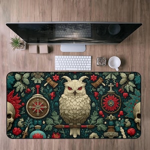 May include: A rectangular desk mat featuring a detailed illustration of a white owl with red eyes, surrounded by floral and skull motifs on a dark teal background. The mat includes decorative elements like axes and shields.