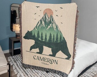 May include: A beige woven throw blanket featuring a mountain and bear design. The design includes a bear silhouette, pine trees, a setting sun, and birds. The name "Cameron" is printed at the bottom.