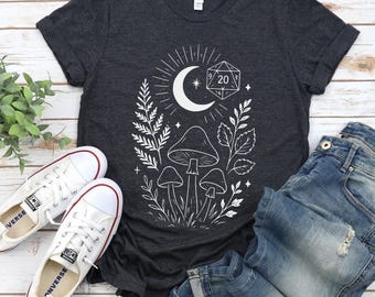 Puede incluir: Camiseta gris oscuro con un gráfico blanco que presenta una luna creciente, setas y un dado de 20 caras. La camiseta se combina con zapatillas blancas y vaqueros azules, creando un look casual.