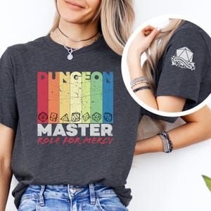 Peut inclure: Un t-shirt gris foncé avec un motif rayé arc-en-ciel qui dit "Dungeon Master Role for Mercy". Le t-shirt a un graphique de dés et les mots "Dungeon Master" sont en blanc. Le t-shirt est porté par une personne.