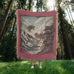 May include: A red and beige woven throw blanket with a Japanese-style illustration of a sea monster in a large wave, with buildings and mountains. The blanket has a fringed edge and is held up in a forest setting.