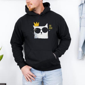 May include: Black hooded sweatshirt with a white cat graphic wearing sunglasses and a gold crown. The cat graphic has the text "Hi Zev!" in yellow. The sweatshirt has a front pocket and a drawstring hood.