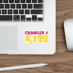 Pode incluir: Um adesivo recortado com o texto CRAWLER # em letras rosa em negrito acima do número 4,122 em grandes dígitos amarelos com um contorno branco. O adesivo está colocado na superfície de um laptop prateado perto de um mouse branco.