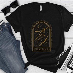 Puede incluir: Camiseta negra con un gr&aacute;fico dorado de una figura corriendo a trav&eacute;s de una tormenta, con el texto "Life Before Death = Strength Before Weakness = Journey Before Destination" y "Stormblessed". Incluye vaqueros azules, gafas de sol negras y zapatillas negras.