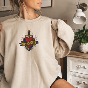 Op de afbeelding: Beige sweatshirt met een hartvormig ontwerp doorboord door een zwaard. Het hart is rood met gele accenten, en het ontwerp bevat een banner met het woord "FIREHEART" in geel. De sweatshirt heeft lange mouwen.