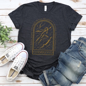 Puede incluir: Camiseta gris oscuro con una ilustraci&oacute;n dorada de una figura en una tormenta, con el texto "Stormblessed". Zapatillas blancas y vaqueros azules est&aacute;n al lado de la camiseta. La camiseta tiene las frases "Life Before Death, Strength Before Weakness, Journey Before Destination."