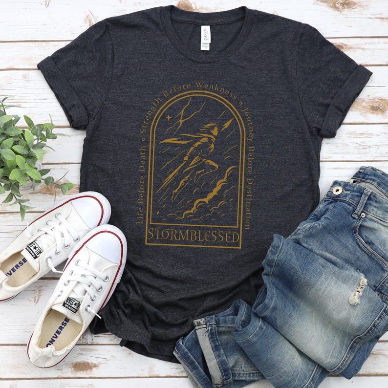 Puede incluir: Camiseta gris oscuro con una ilustraci&oacute;n dorada de una figura en una tormenta, con el texto "Stormblessed". Zapatillas blancas y vaqueros azules est&aacute;n al lado de la camiseta. La camiseta tiene las frases "Life Before Death, Strength Before Weakness, Journey Before Destination."