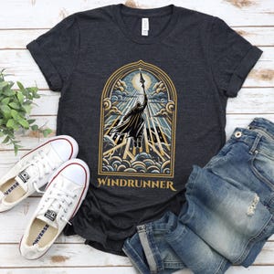 May include: Dark gray t-shirt with a graphic of a figure reaching for a spear, framed by an archway with the word "WINDRUNNER" below. White sneakers and blue jeans are next to the shirt.