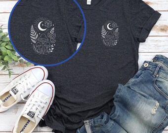 Puede incluir: Camiseta gris oscuro con un diseño blanco de una luna creciente, setas y follaje. El diseño incluye un dado de 20 caras. La imagen incluye zapatillas blancas y vaqueros azules.