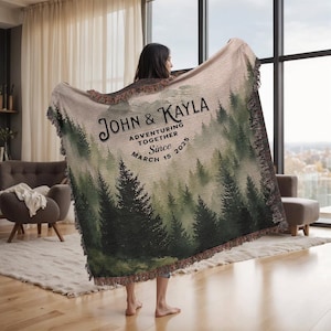 May include: A woven blanket with a forest design, showing evergreen trees and a misty backdrop. The blanket displays the text "John & Kayla Adventuring Together Since March 15 2025". It has a fringed border and is held up by a person.