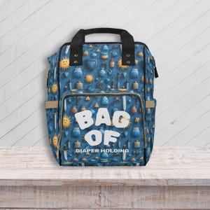 Può includere: Borsa per pannolini blu con un motivo ripetuto di soli, spade e fiale di pozioni. La borsa ha le parole "BAG OF DIAPER HOLDING" in lettere bianche. Ha un manico e finiture nere.