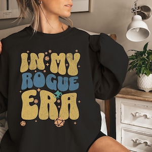 May include: Black sweatshirt with the text "IN MY ROGUE ERA" in yellow and blue, with dice and star graphics. The design has a retro, vintage style. The sweatshirt is a casual garment.