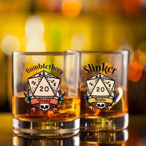 Könnte beinhalten: Zwei klare Glasbecher mit brauner Flüssigkeit. Jeder Becher hat einen bunten Aufkleber mit einem 20-seitigen Würfel, Blumen und einem Totenkopf. Der Text auf den Aufklebern lautet "Bumblethorn Druid" und "Slinker Rogue".