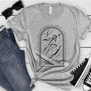 Puede incluir: Una camiseta gris con un gr&aacute;fico negro de una figura corriendo a trav&eacute;s de una tormenta, con el texto "Life Before Death, Strength Before Weakness, Journey Before Destination" y "Stormblessed". La camiseta se combina con vaqueros azules y zapatillas negras.