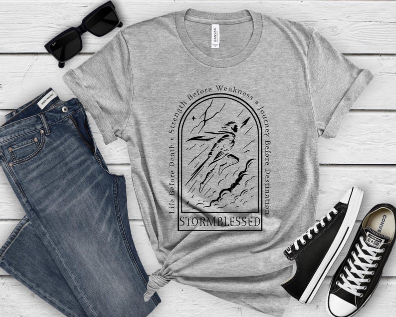 Puede incluir: Una camiseta gris con un gr&aacute;fico negro de una figura corriendo a trav&eacute;s de una tormenta, con el texto "Life Before Death, Strength Before Weakness, Journey Before Destination" y "Stormblessed". La camiseta se combina con vaqueros azules y zapatillas negras.