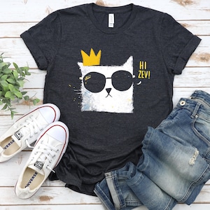 May include: Dark grey t-shirt with a white cat wearing sunglasses and a yellow crown. The shirt has the text "Hi Zev!" and is paired with white trainers and blue jeans. The shirt is made of a soft material.