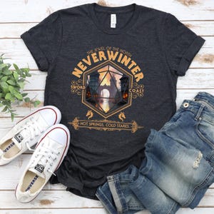 May include: A dark gray t-shirt with a graphic design featuring the text "Neverwinter" in a gold font. The graphic also includes a stylized image of a city with a large, gold-colored dice in the center. The text "The Jewel of the North" appears above the graphic, and the text "Sword Coast" appears to the right of the graphic. The text "Hot Springs, Cold Stares" appears below the graphic.