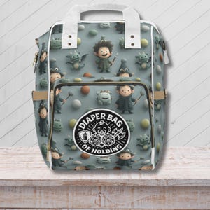 Può includere: Una borsa per pannolini grigia con manico e finiture bianche, con un motivo ripetuto di personaggi dei cartoni animati e la scritta "DIAPER BAG OF HOLDING". La borsa ha una tasca frontale e una tasca laterale.