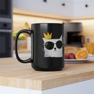 May include: Black ceramic mug featuring a white cat illustration wearing sunglasses and a gold crown. The cat design includes the text "Hi Zzz". The mug has a large handle and is displayed on a wooden surface.