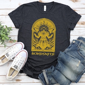 May include: Dark gray t-shirt with a gold graphic of a figure in an archway, with the word "BONDSMITH" below. The shirt is paired with white sneakers and blue jeans on a light wood surface.