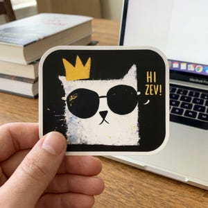 Könnte beinhalten: Ein quadratischer Aufkleber mit einer weißen Katze mit Sonnenbrille und goldener Krone auf schwarzem Hintergrund. Der Aufkleber trägt den weißen Text "HI ZEV!". Die Katze hat ein strukturiertes, gemaltes Aussehen.