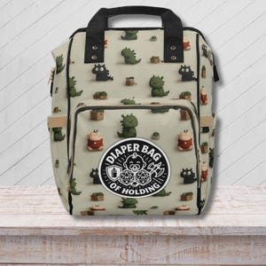 Può includere: Una borsa per pannolini beige con un motivo ripetuto di dinosauri dei cartoni animati, forzieri e figure di bambini. La borsa ha un manico nero e una tasca frontale con la scritta "Diaper Bag of Holding" in un emblema circolare.