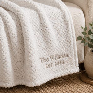 May include: A white textured throw blanket featuring a popcorn-style knit pattern. The lower corner is embroidered with the text "The Wilsons" above "EST. 2026" in a light brown thread, providing a clear and legible personalized detail on the fabric.