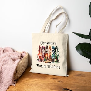 May include: A white canvas tote bag with the text "Christina's Bag of Holding" printed on the front. The bag features a colourful illustration of a wizard's bag with various magical items, including a sword, a potion bottle, a book, and a dragon.