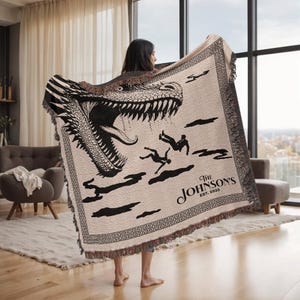 May include: A beige woven throw blanket featuring a black and white dinosaur graphic with the text "The Johnson's EST. 2025". The blanket has a fringed edge and a geometric border design.