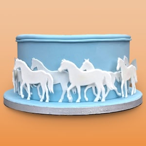 Puede incluir: Un pastel azul claro decorado con siluetas de caballos blancos. El pastel, de forma redonda, se asienta sobre una base plateada. Los caballos, dispuestos alrededor de la base, crean un efecto de carrusel.