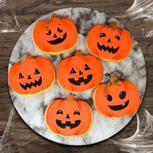 May include: Six orange pumpkin-shaped cookies with black icing jack-o'-lantern faces on a white marble plate.