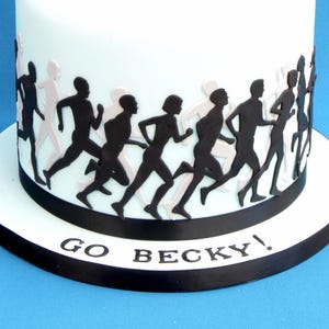 May include: A white cylindrical cake decorated with a band of black and light-coloured silhouettes of people running. The base of the cake features a black ribbon and the text GO BECKY! printed in black capital letters on the white cake board.