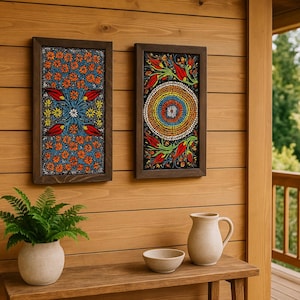 Może przedstawiać: Dwa oprawione obrazy ścienne ze skomplikowanymi wzorami kwiatowymi. Jeden przedstawia pomarańczowe kwiaty na niebieskim tle, drugi okrągły wzór z czerwonymi, żółtymi i niebieskimi elementami. Ramy są ciemnobrązowe.