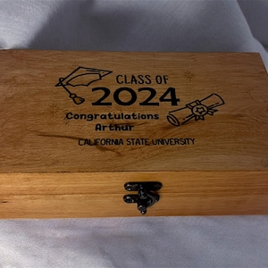 May include: A wooden box with a hinged lid and a latch. The top of the box is engraved with the text "Class of 2024", "Congratulations Arthur", and "California State University". There are also illustrations of a graduation cap and a diploma.