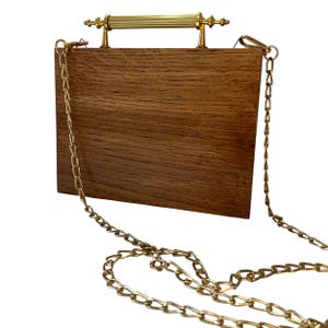 May include: A rectangular wooden handbag with a gold-colored chain strap and handle. The bag is made of brown wood with a visible grain pattern. The handle is a gold-colored bar with decorative ends.